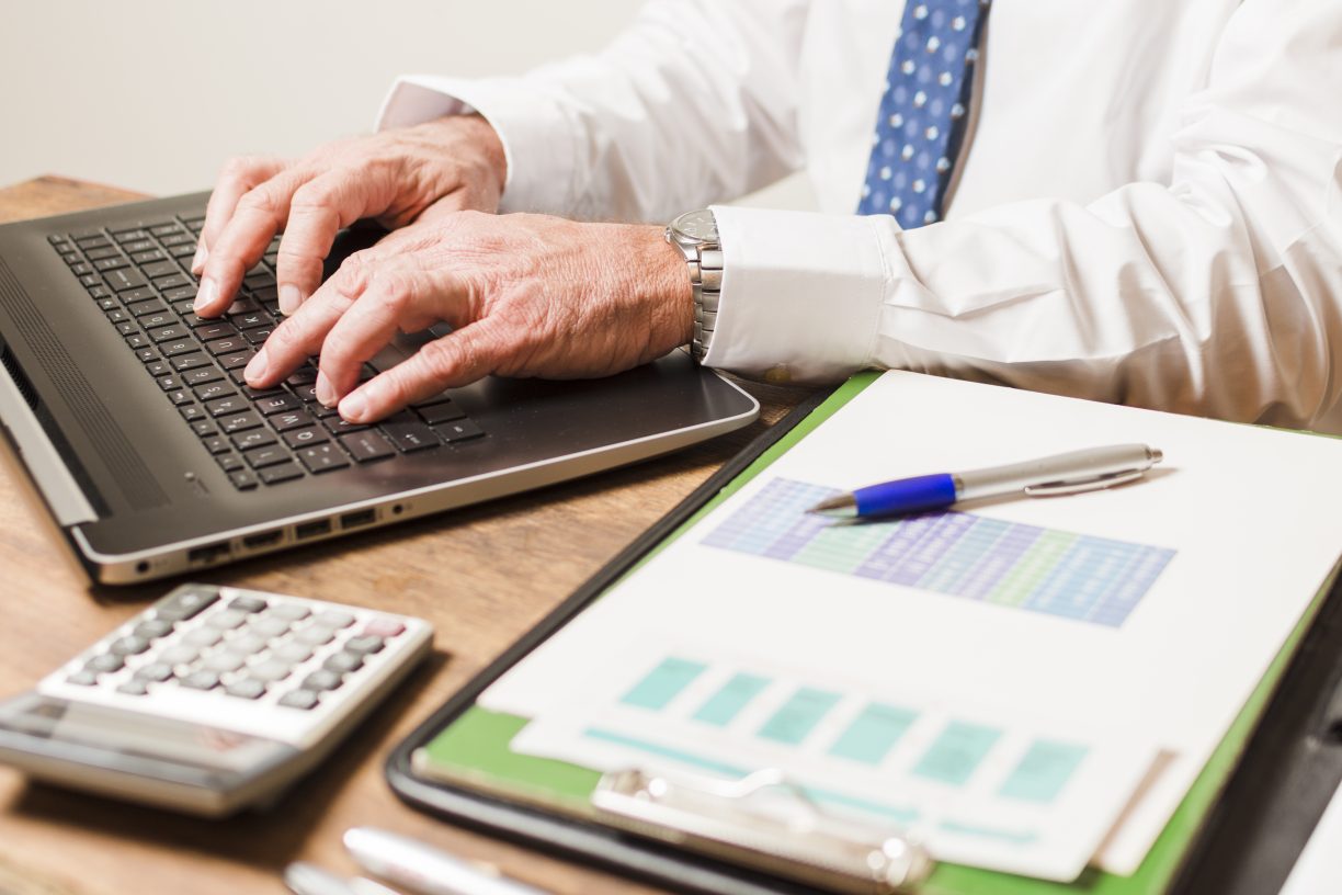 a man performs a payroll health check at his laptop, with paperwork and a calculator on the desk near him